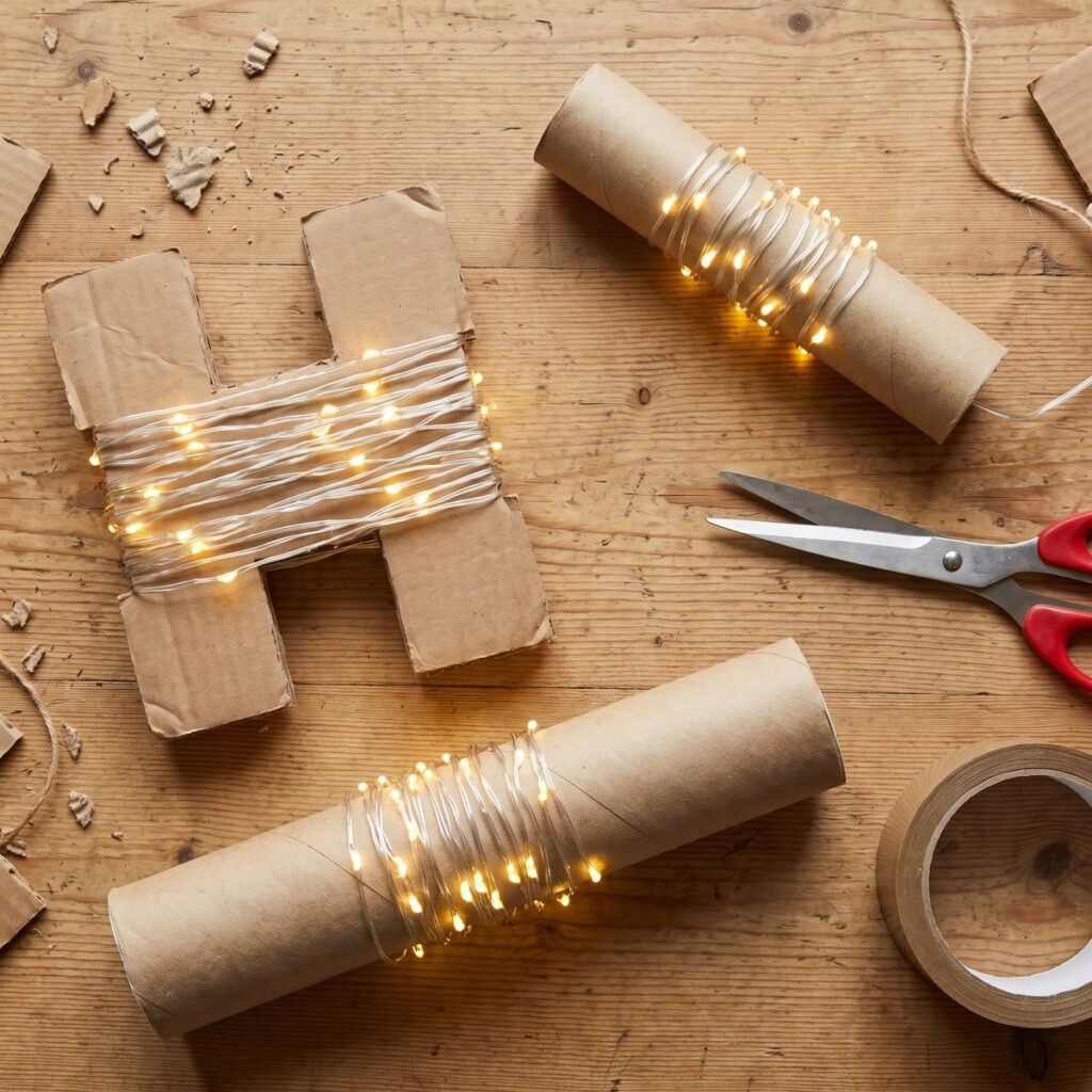 Warm white fairy lights wound neatly around cardboard H shapes cut from a delivery box and around a kitchen roll tube, with scissors and parcel tape on a wooden table